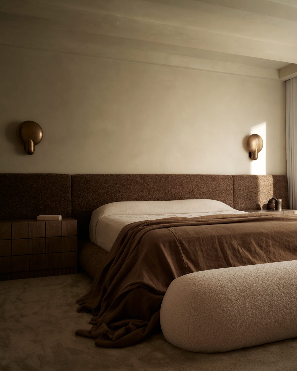 A channel-upholstered headboard wall in warm brown with brass sconces