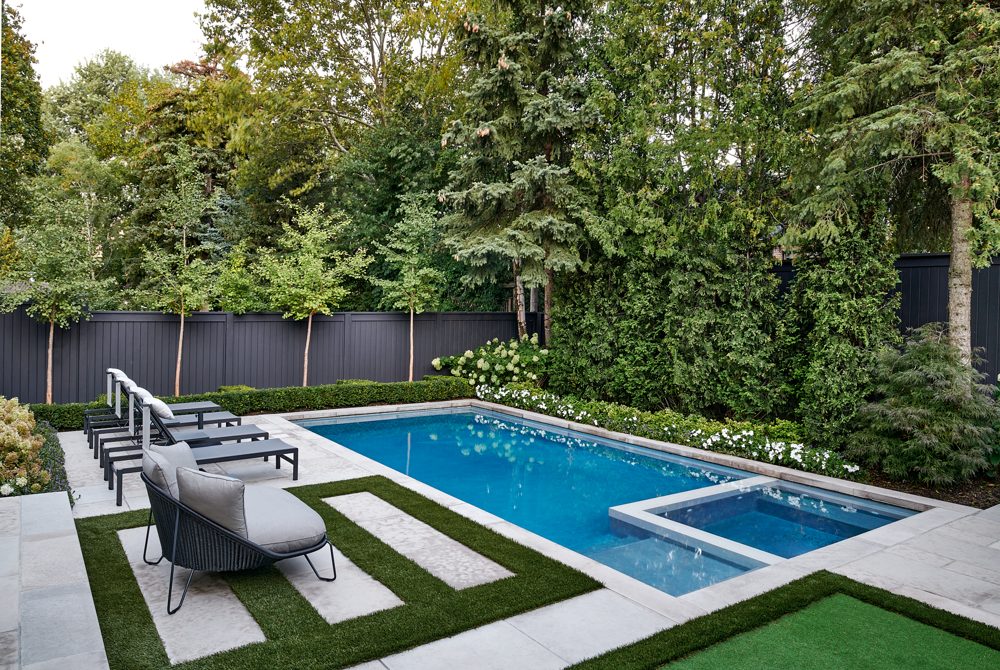 A garden with a pool, a row of loungers and a black fence under tall trees