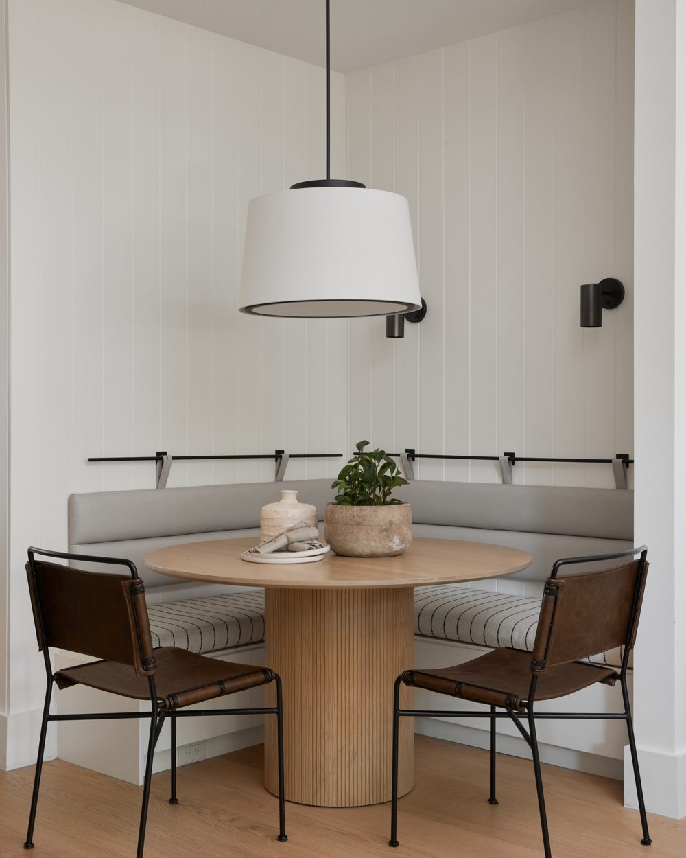 A grey banquette around a fluted oak pedestal table under a white drum pendant